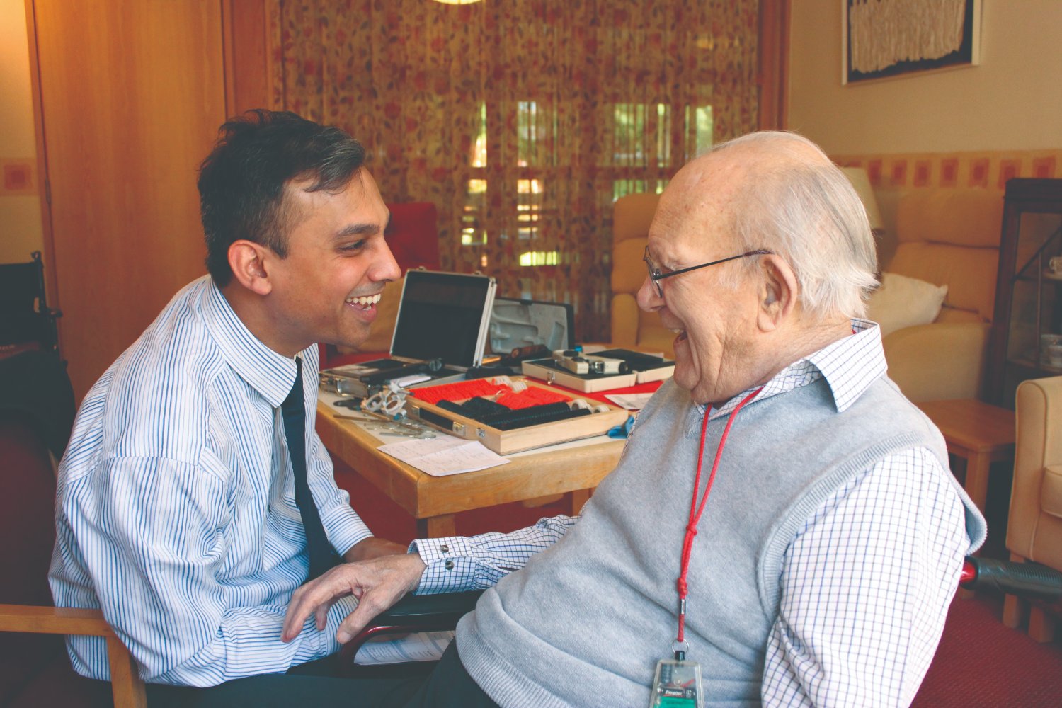 An Opticall optician sharing a warm moment with an older gentleman during a home eye test, trial frames laid out on the table