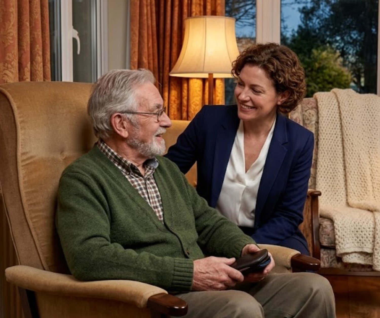 An Opticall optician sitting beside an older gentleman in his armchair at home, talking warmly as he holds his glasses